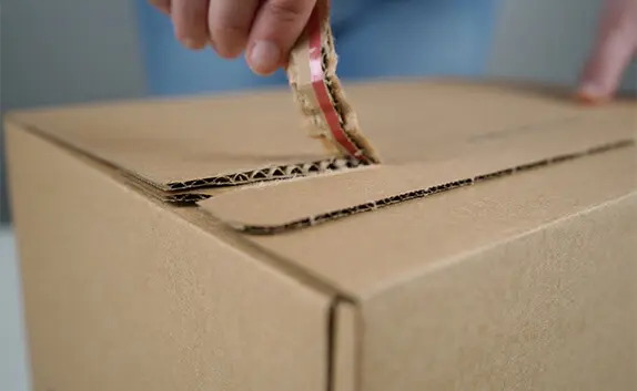 Nahaufnahme, wie eine Person einen Karton an einem Aufreißfaden öffnet.