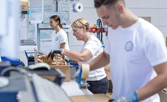 Logistikmitarbeiter stehen an Packtischen und verpacken Produkte in Paketen.