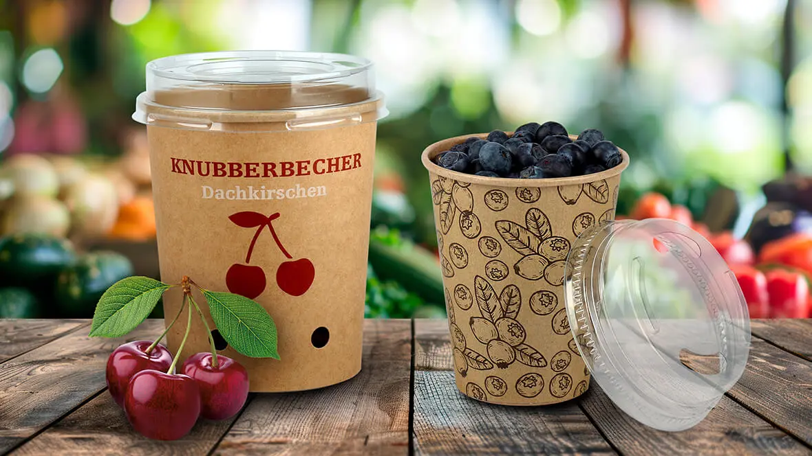 Auf einem Tisch stehen Pappbecher mit Kunststoff-Deckel für Kirschen und Blaubeeren