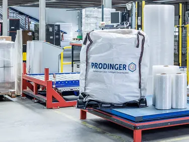 Ein Big Bag von PRODINGER steht bei einer Stretchmaschine mit mehreren Rollen Stretchfolie in einer Logistikhalle.