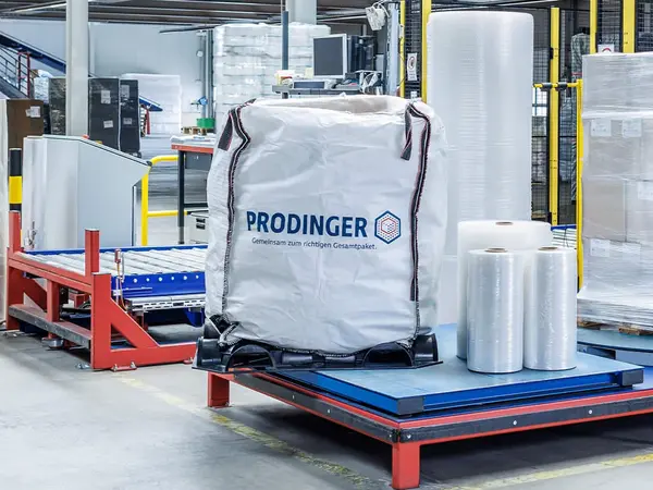 Ein Big Bag von PRODINGER steht bei einer Stretchmaschine mit mehreren Rollen Stretchfolie in einer Logistikhalle.
