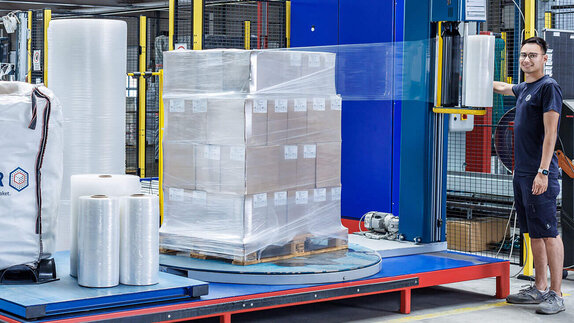 Lagerist bedient eine Stretchmaschine, die eine Palette mit Paketen in Stretchfolie umwickelt.