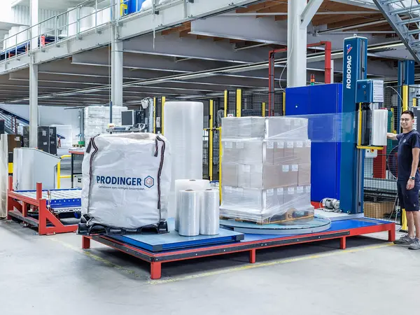 In einer Lager wickelt ein Logistikmitarbeiter eine Palette mit einer Stretchmaschine ein. Weiter Folienprodukte stehen im Hintergrund.