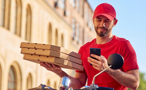 Ein Pizzalieferant sitzt auf einem Roller und hält mehrere Pizzen in der Hand.